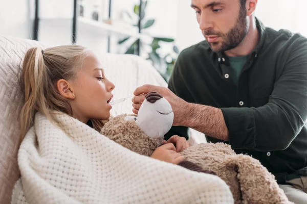 Atento padre dando medicamentos a la hija enferma sosteniendo osito de peluche - foto de stock