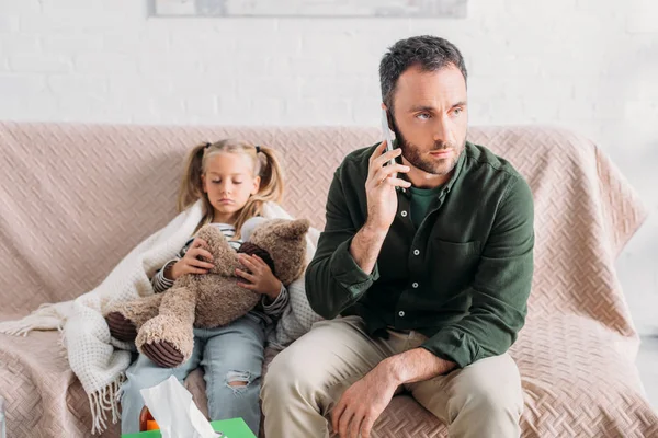 Homme sérieux parlant sur smartphone tout en étant assis près de fille malade — Photo de stock