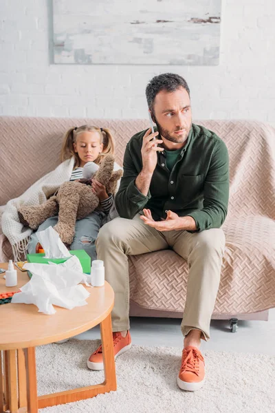 Серьезный отец разговаривает по смартфону, сидя рядом с больной дочерью и столом с лекарствами — стоковое фото