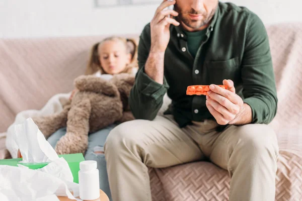 Vista recortada del hombre hablando en el teléfono inteligente y sosteniendo ampolla de píldoras mientras está sentado cerca de hija enferma - foto de stock