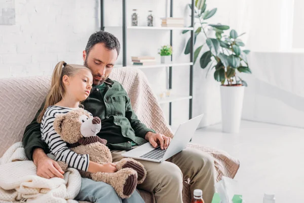 Molesto, niño enfermo sosteniendo oso de peluche mientras está sentado en el sofá cerca de padre - foto de stock