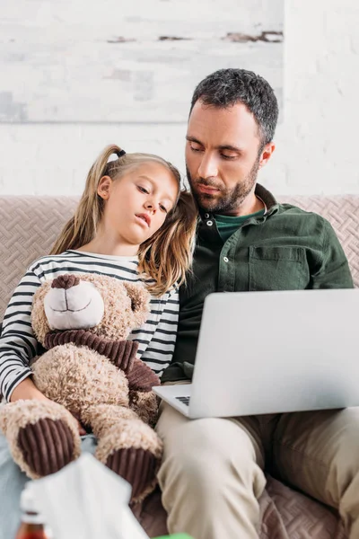 Diseased kid using laptop with father while sitting on sofa — Stock Photo