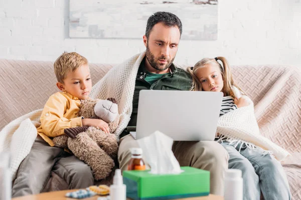 Niños enfermos sentados en el sofá cerca de padre enfermo usando el ordenador portátil - foto de stock
