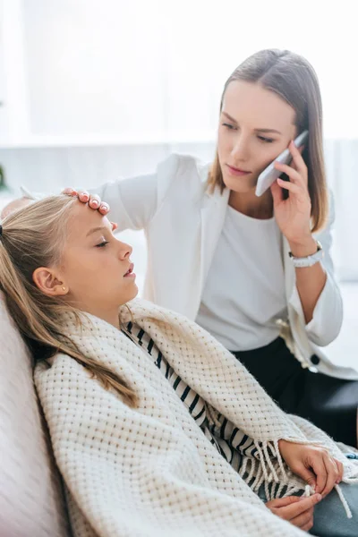 Madre preocupada tocando la frente de la hija enferma mientras habla en el teléfono inteligente - foto de stock