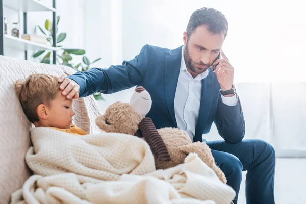 Padre preoccupato toccare la fronte del figlio malato mentre parla su smartphone — Foto stock