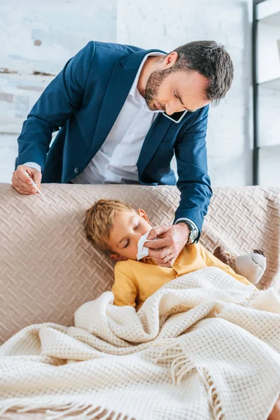 Père attentionné essuyant nez de fils malade tout en tenant thermomètre et parler sur smartphone — Photo de stock