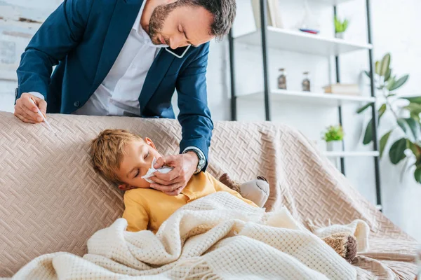 Padre attento asciugare il naso del figlio malato mentre tiene il termometro e parla sullo smartphone — Foto stock