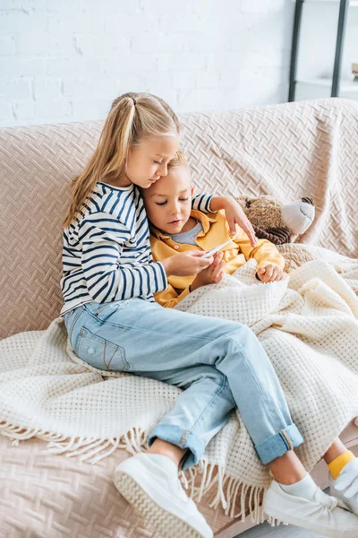 Atenta hermana mirando el termómetro mientras abraza al hermano enfermo - foto de stock