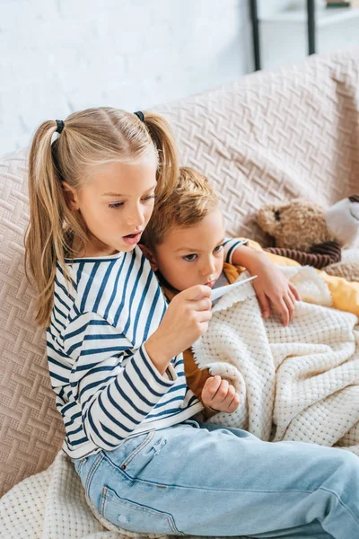 Hermana sorprendida mirando el termómetro mientras abrazaba al hermano enfermo - foto de stock