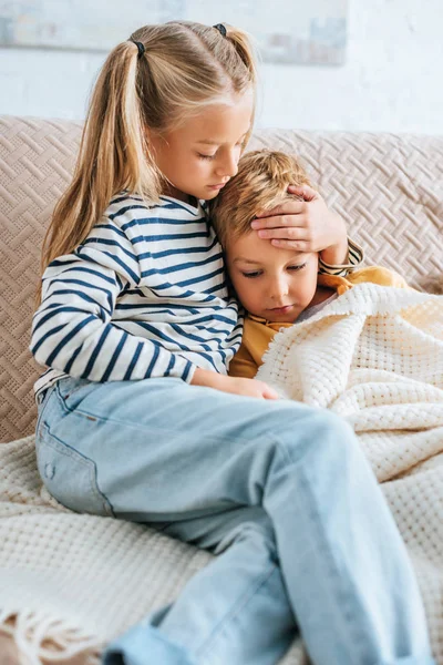 Attentive sister touching forehead of ill brother while sitting on sofa together — Stock Photo