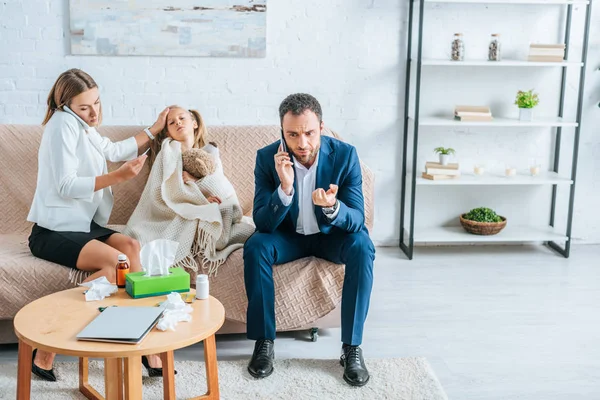 Padres preocupados hablando en teléfonos inteligentes mientras están sentados cerca de hija enferma cubierta con manta - foto de stock