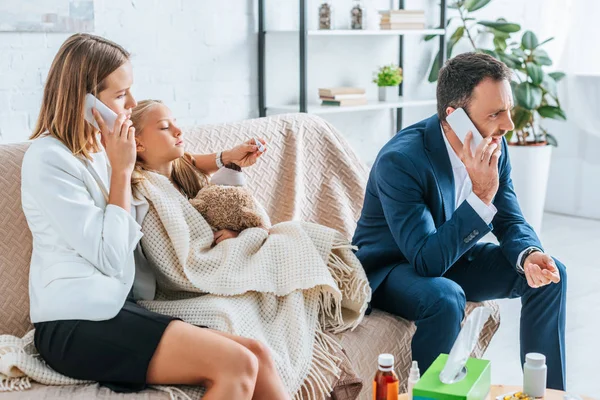 Worried parents talking on smartphones while sitting near diseased daughter covered with blanket — Stock Photo