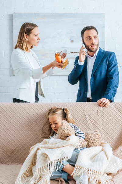 Madre preocupada sosteniendo medicamentos y el padre hablando en el teléfono inteligente cerca de hija enferma sentada en el sofá debajo de la manta - foto de stock