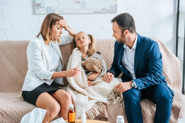 Besorgte Mutter berührt Stirn und blickt auf Thermometer, während sie neben kranker Tochter und Ehemann sitzt — Stockfoto