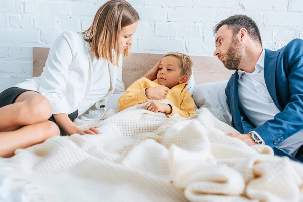 Preocupado marido y mujer mirando enfermo hijo acostado en la cama - foto de stock