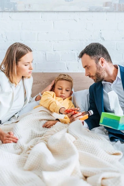 Worried father holding cough syrup and mother touching head of diseased son lying in bed — Stock Photo