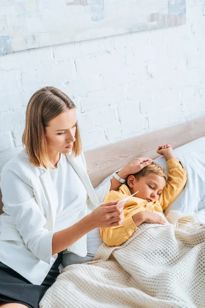 Mère inquiète regardant thermomètre tout en touchant la tête du fils malade — Photo de stock