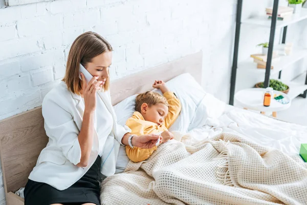 Mujer preocupada mirando el termómetro y hablando en el teléfono inteligente mientras está sentado cerca de hijo enfermo - foto de stock