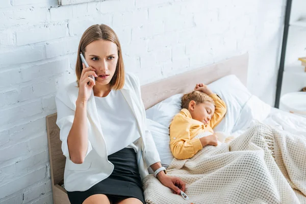 Besorgte Frau spricht auf Smartphone, während sie in der Nähe ihres kranken Sohnes sitzt — Stockfoto