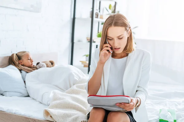 Mujer preocupada sosteniendo botiquín de primeros auxilios y hablando en el teléfono inteligente mientras está sentada cerca de hija enferma - foto de stock