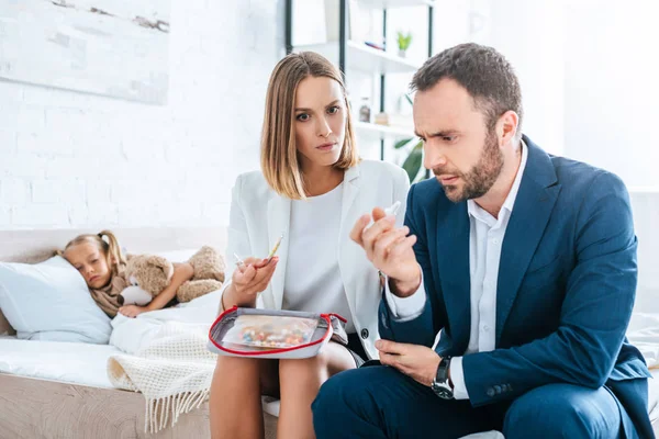 Entmutigte Eltern in offizieller Kleidung sitzen auf Sofa mit Verbandskasten neben kranker Tochter im Bett liegend — Stockfoto