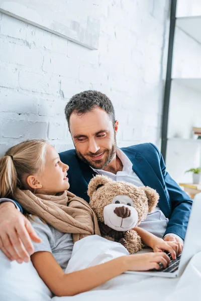 Krankes Kind schaut lächelnden Vater beim gemeinsamen Benutzen des Laptops an — Stockfoto