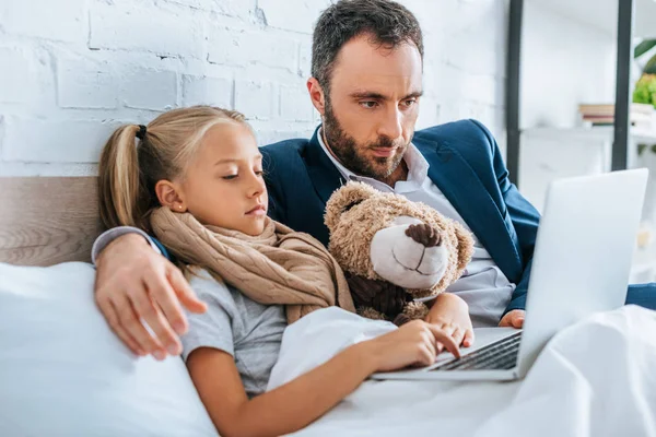 Enfant malade utilisant un ordinateur portable avec son père alors qu'il était couché au lit — Photo de stock