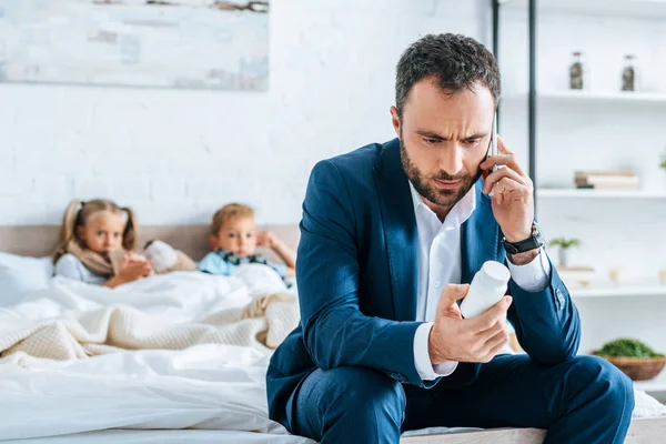 Hombre molesto hablando en el teléfono inteligente y sosteniendo contenedor con pastillas mientras está sentado en la cama cerca de niños enfermos - foto de stock