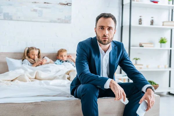 Selective focus of man in formal wear holding container with pills and smartphone while sitting on bed near diseased children — Stock Photo