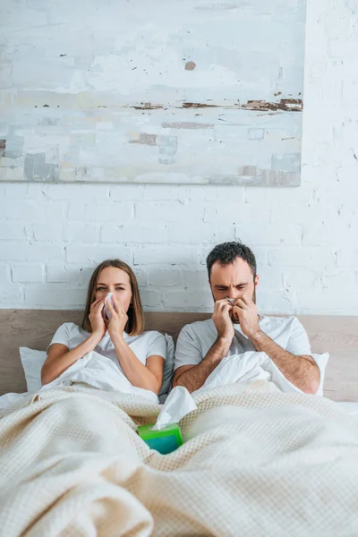 Marido y mujer enfermos estornudando mientras yacen juntos en la cama - foto de stock