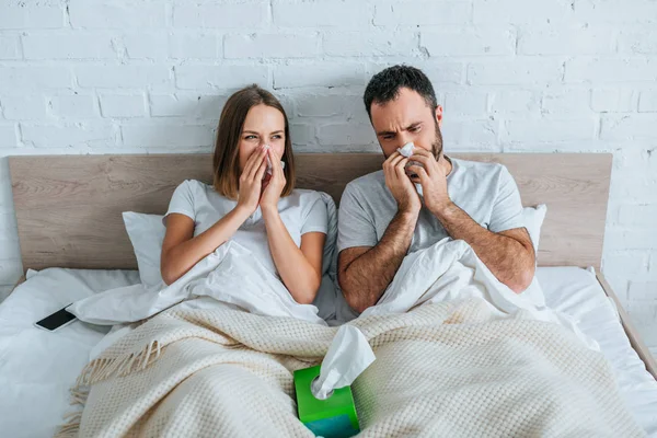 Marido y mujer enfermos estornudando mientras yacen juntos en la cama - foto de stock