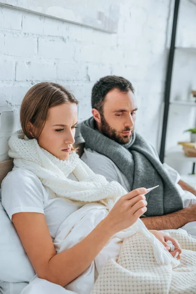 Kranke Frau blickt neben krankem Ehemann auf Thermometer — Stockfoto