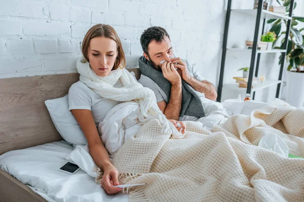 Sick woman looking at thermometer near ill husband sneezing in napkin — Stock Photo