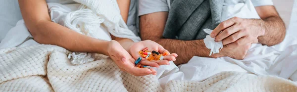 Vista recortada de la mujer enferma sosteniendo un puñado de pastillas mientras está acostado en la cama cerca de marido enfermo, tiro panorámico - foto de stock