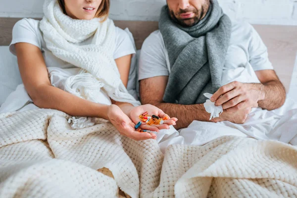 Vista cortada de mulher doente segurando punhado de pílulas enquanto sentado na cama perto do marido doente — Fotografia de Stock