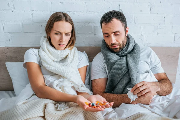 Mujer enferma sosteniendo un puñado de pastillas mientras está sentada en la cama cerca del marido enfermo - foto de stock