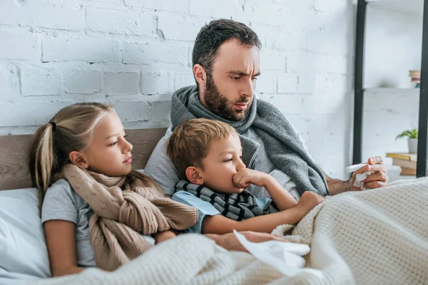Засмучений батько дивиться на термометр, лежачи в ліжку з хворими дітьми — стокове фото