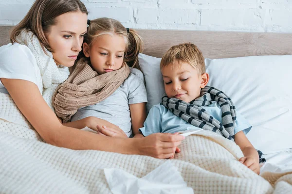 Madre enferma mostrando termómetro a los niños enfermos mientras están acostados juntos en la cama - foto de stock