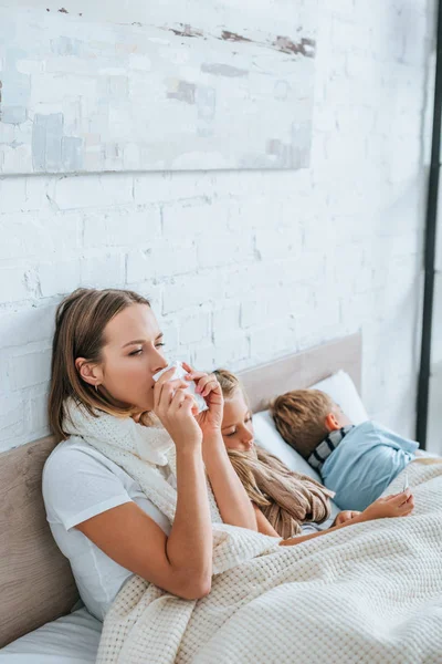 Больная женщина чихает в салфетку, сидя в постели рядом с больными детьми — стоковое фото