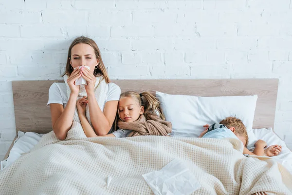 Больная женщина чихает в салфетку, сидя в постели рядом с больными детьми — стоковое фото