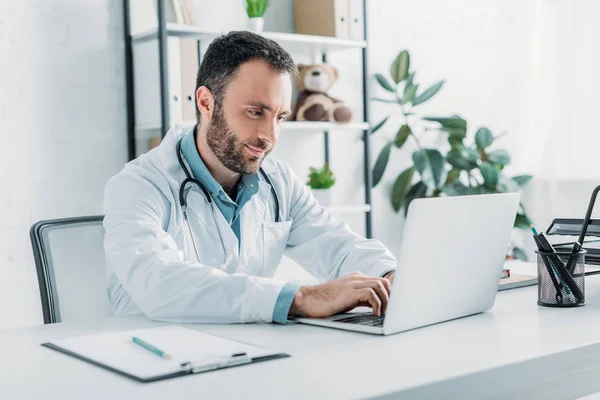 Doctor positivo sentado en el lugar de trabajo y usando el ordenador portátil - foto de stock