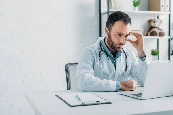 Medico premuroso guardando computer portatile mentre seduto sul posto di lavoro — Foto stock