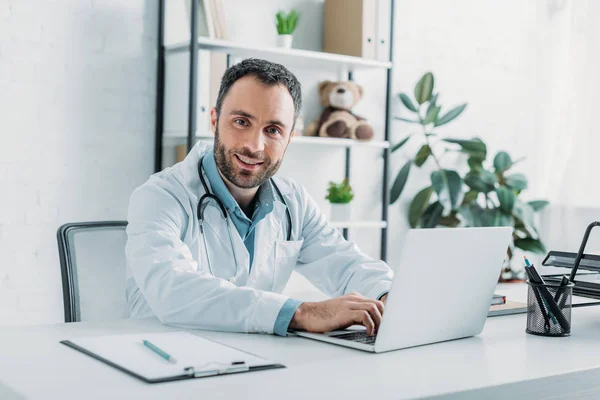 Sonriente médico mirando a la cámara mientras usa el ordenador portátil - foto de stock