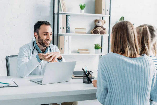 Medico sorridente guardando la madre e il bambino seduto alla scrivania — Foto stock