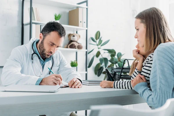 Pediatra serio escribir prescripción cerca de la madre y la hija - foto de stock