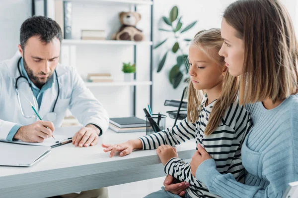 Diagnóstico serio de la escritura del doctor cerca de madre y de hija atentas - foto de stock