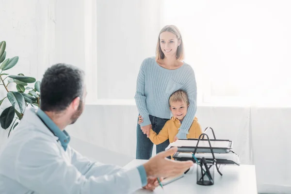 Lächelnde Mutter umarmt fröhlichen Sohn beim Blick auf Kinderarzt im Krankenhaus — Stockfoto