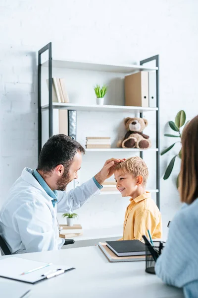 Pediatra examinando sonriente chico cerca de madre sentado en escritorio - foto de stock