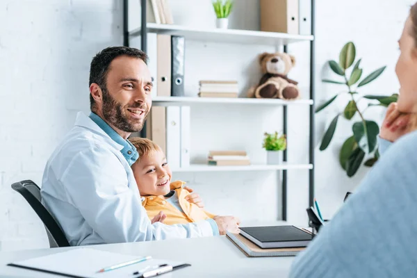 Souriant pédiatre étreignant garçon tout en regardant mère assis au bureau — Photo de stock