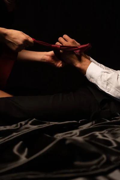 Обрезанный вид женщины, связывающей руки мужчины с галстуком на черной кровати — стоковое фото